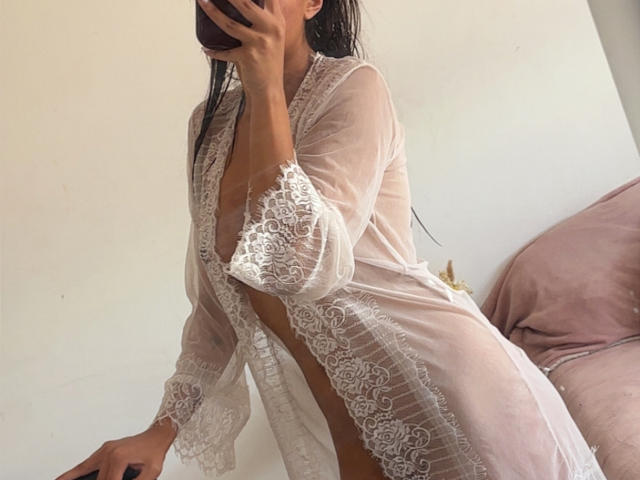 DianaFernandezz indossa un abito bianco in pizzo trasparente, parzialmente aperto per rivelare il petto e l'addome. La sua mano copre la bocca, lunghi capelli scuri le ricadono sulle spalle. Un cuscino rosa chiaro sullo sfondo..