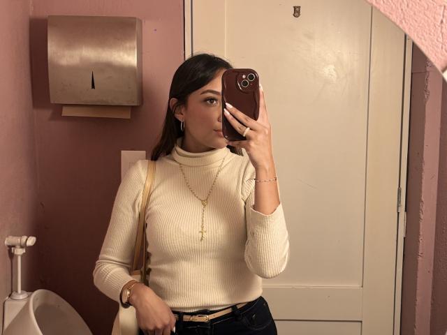 DianaFernandezz, in a pink bathroom, takes a mirror selfie with her phone. She wears a tight, cream ribbed sweater, dark pants, gold necklace, and bracelet. She's holding her phone in front of her face.