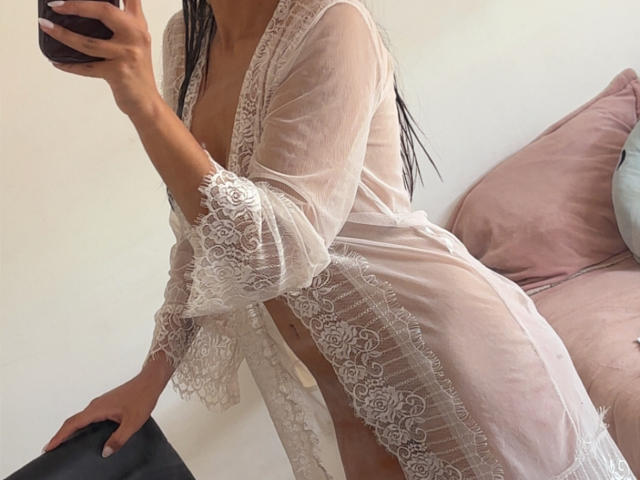 Photograph of DianaFernandez with long braids, wearing a sheer white lace robe, exposing her breasts and midriff. She holds a phone in her right hand, leaning forward, showing a hint of her cock. Pink pillow in the background.