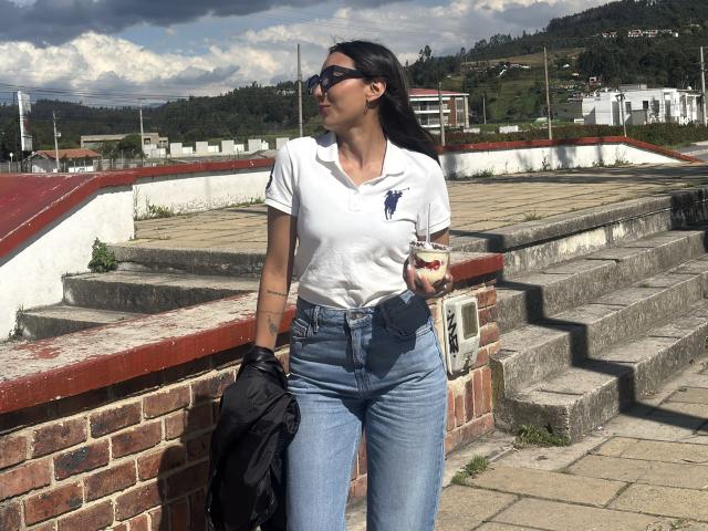 DianaFernandezz leans against a brick wall, wearing a tight white t-shirt and high-waisted blue jeans, holding a black jacket. She's gazing off to the side, under a cloudy sky.