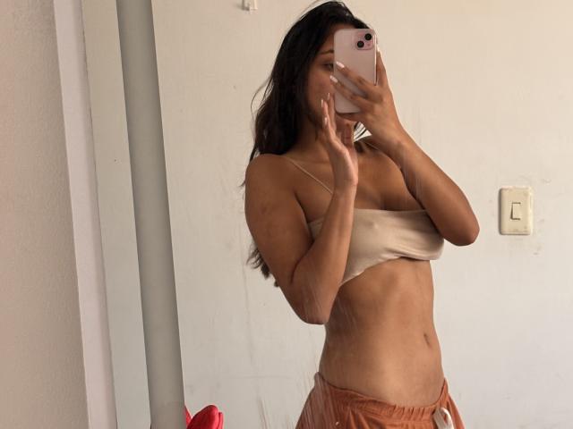 Fotografie von Diana Fernandez, die ein Selfie vor einem Spiegel macht. Sie trägt ein beiges Bandeau-Top und braune Hosen und hält ihr Gesicht mit ihrem Handy bedeckt. Ihre straffen Bauchmuskeln sind sichtbar, und im Hintergrund befindet sich ein rotes Objekt..