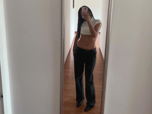 DianaFernandezz steht vor einem Spiegel und macht ein Selfie. Sie trägt ein weißes Crop-Top, das ihren Bauch freilegt, und schwarze Hosen. Langes, schwarzes Haar bedeckt einen Teil ihres Gesichts..