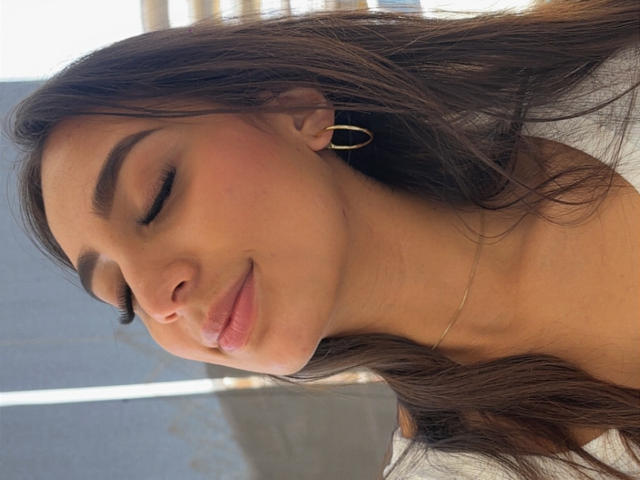 Primer plano de DianaFernandez con los ojos cerrados, una sonrisa suave y las mejillas sonrojadas. Cabello largo y castaño, aros dorados y un collar delgado. La luz del sol resalta su piel. Absolutamente precioso, ambiente relajado..