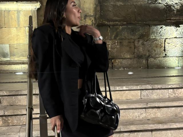 Fotografie von Diana Fernandez, die auf einer schlecht beleuchteten Steintreppe steht, an einem Geländer lehnt und einen schwarzen Mantel trägt. mit einer schwarzen Handtasche an ihrer rechten Seite, die eine sexuelle Spannung ausstrahlte..