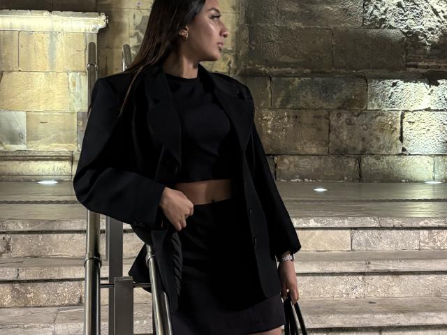 Fotografie von Diana Fernandez, die vor einer Steinmauer steht, ein schwarzes Crop-Top und eine Jacke trägt und eine schwarze Tasche hält. Lange Zöpfe, ernster Gesichtsausdruck, schwach beleuchtet, Schatten auf steinernen Stufen..