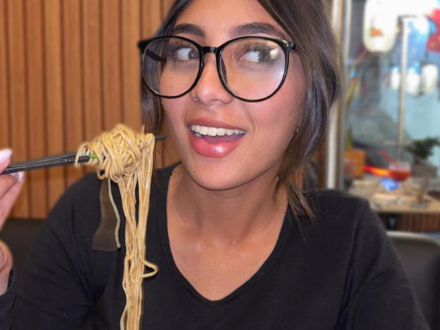 DianaFernandezz, con occhiali dalla montatura nera e spessa, tiene una forchetta con spaghetti aggrovigliati vicino alla bocca, sorridendo con le labbra lucide. labbra rosa, indossa una maglietta nera, sfondo con pannelli di legno, leggermente sfocato.