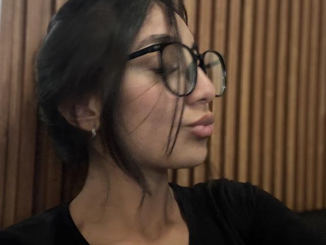 Photograph of DianaFernandezz with dark hair, black-rimmed glasses, and a pouty expression. She's wearing a black shirt, wooden background, and small earrings. Fucking sexy, glasses add nerdy charm.