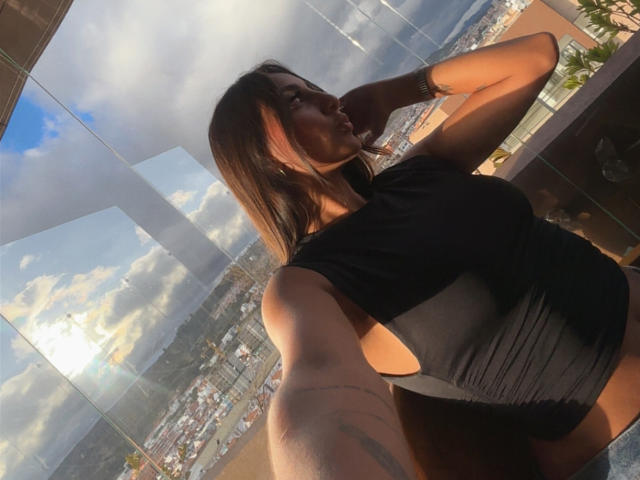 DianaFernandezz takes a selfie in a sunlit room with large glass windows. She's wearing a tight black top, highlighting her large breasts, and has a tattoo on her right arm. Fucking sexy.