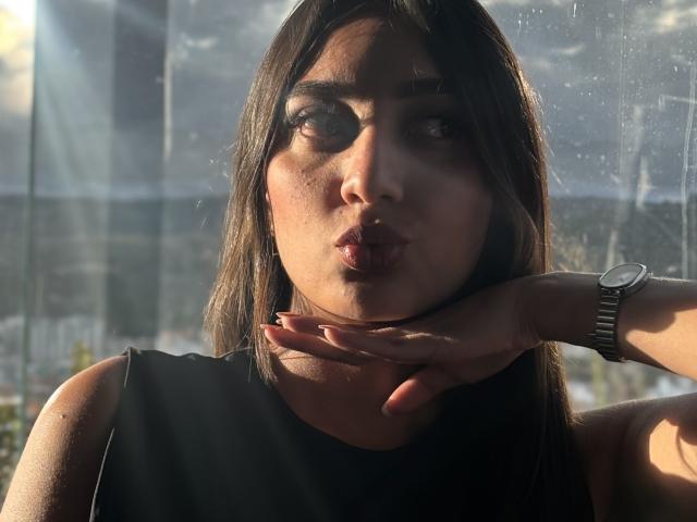 Photographie de Diana Fernandez, une femme aux longs cheveux noirs, aux lèvres pincées et au regard intense, portant une robe noire sans manches. vue de dessus, main reposant sur le cou, la lumière du soleil projetant des ombres, montre au poignet..