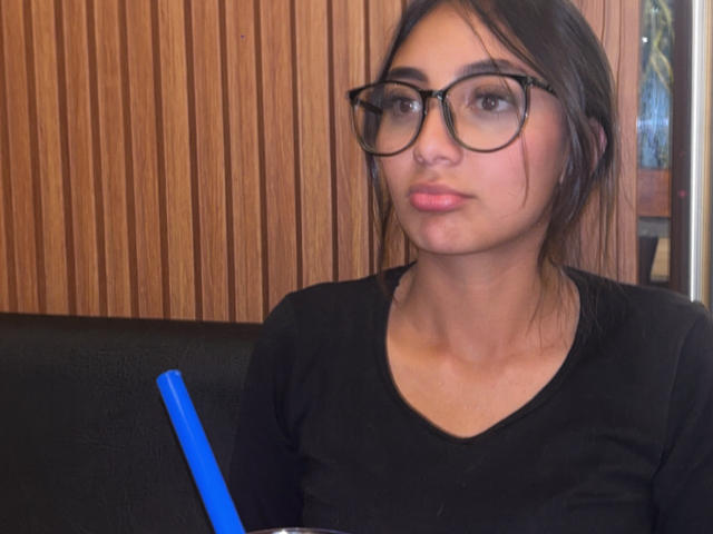 DianaFernandezz, una donna con capelli scuri e occhiali, che indossa una maglietta nera, seduta davanti a un laptop blu, con un pannello di legno sullo sfondo. sfondo, espressione seria, labbra rosa leggermente dischiuse, foto casuale.