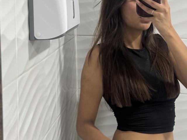 Fotografia de DianaFernandezz com cabelos castanhos longos, vestindo uma cropped preta justa, segurando um telefone no rosto. Cobrindo a boca e o nariz, em um banheiro com azulejos brancos. Muito atraente..