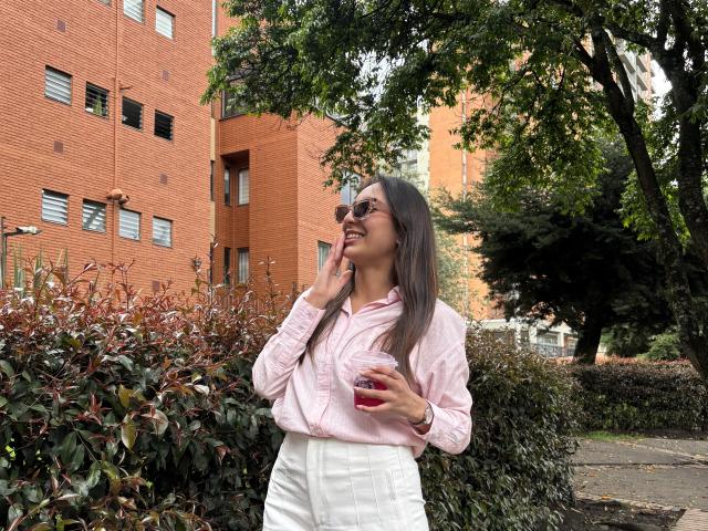 DianaFernandezz se tient dans un environnement urbain ensoleillé, vêtue d'une chemise rose à manches longues et d'un pantalon blanc, et tient un objet rouge. Elle est appuyée contre un buisson, avec un bâtiment en briques en arrière-plan..