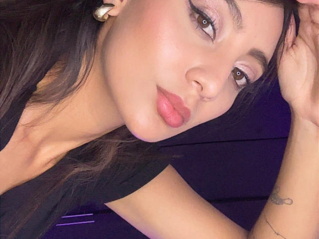 Primer plano de DianaFernandez con cabello oscuro, ojos marrones y labios rosados. Está acostada, con la cabeza inclinada y la mano en el pelo. Lleva una camiseta negra, aretes blancos y un tatuaje pequeño en la muñeca. Apariencia sexy y seductora.