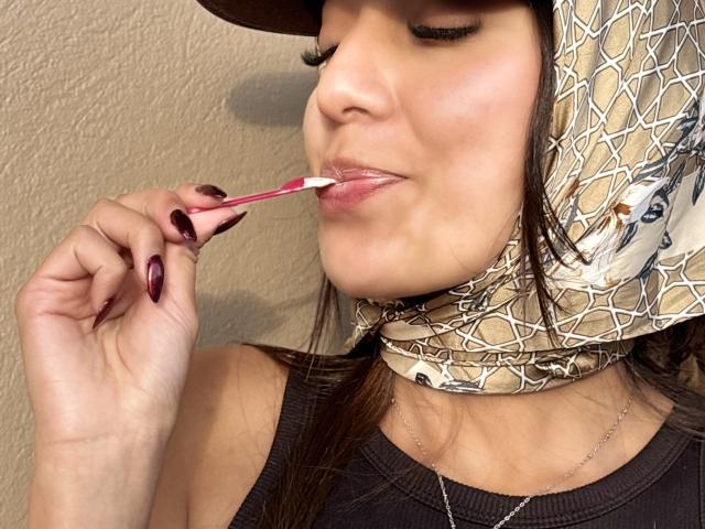 Close-up de DianaFernandez com uma mão segurando um batom rosa, aproximado de seus lábios ligeiramente entreabertos. Ela veste uma camiseta preta e um adorno de cabeça branco com um padrão rachado. As unhas dela são roxas escuras..