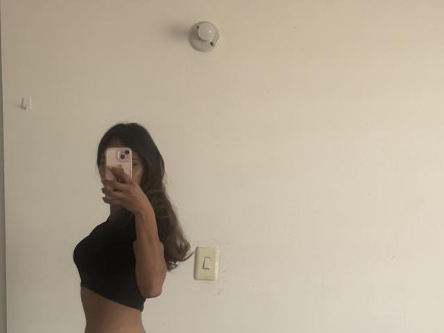 DianaFernandezz prend un autoportrait, tenant un téléphone dans sa main droite, montrant son profil. Elle porte un crop top noir, qui met en valeur sa petite poitrine et sa silhouette mince. Mur blanc, interrupteur et détecteur de fumée en arrière-plan..