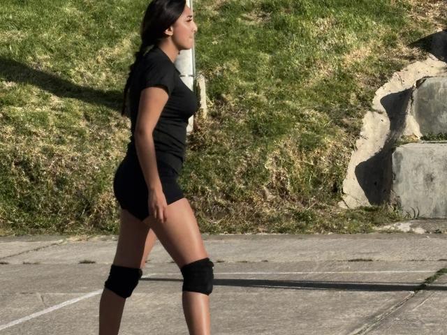 DianaFernandezz marchant, vue de profil, robe noire courte, protège-tibias noirs, cheveux foncés, peau bronzée, fond avec de l'herbe. la lumière du soleil projetant des ombres, environnement extérieur, démarche assurée, espace visible entre les cuisses, tension sexuelle..