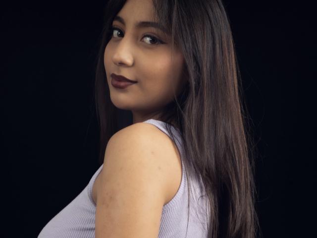 Retrato digital de DianaFernandezz, mirando por encima del hombro con una expresión sensual. Ella tiene el pelo largo, lacio y negro, lápiz labial oscuro, y lleva una camiseta blanca sin mangas. Fondo negro. Tensión sexual..