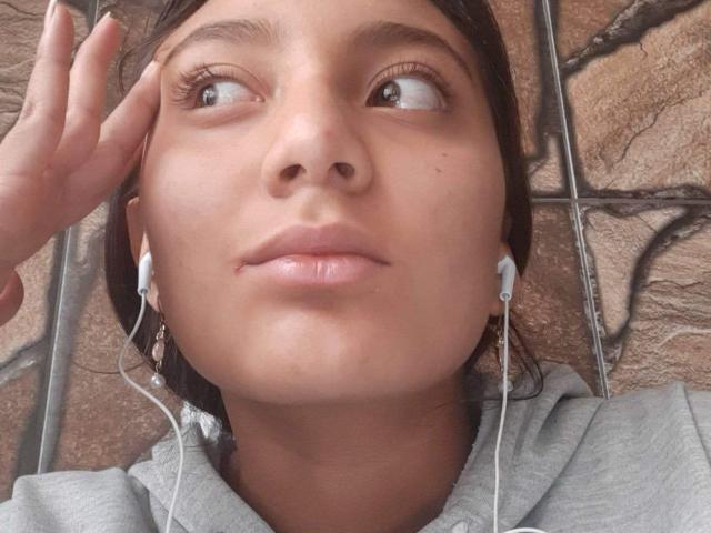 Close-up of DianaFernandez lying on a tiled surface, looking up with a slight gaze, hand touching her temple, wearing a grey hoodie, white earbuds in ears, natural light.