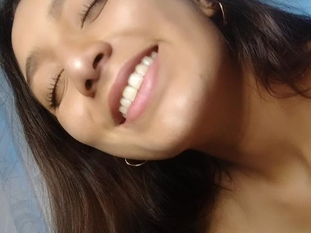 Foto em close-up de DianaFernandezz, com os olhos fechados, sorrindo amplamente, mostrando os dentes. Seus cabelos escuros emolduram seu rosto, e a luz do sol realça sua pele bronzeada. Ela usa pequenas argolas nos ouvidos. Um sorriso incrivelmente lindo..