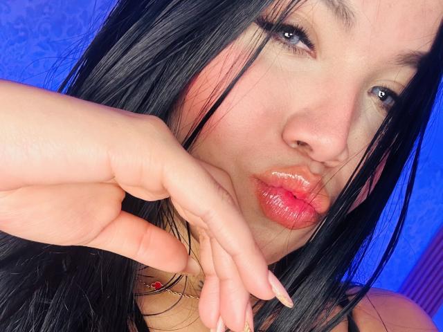Close-up photo of MiaDenver with glossy red lips, black hair, and blue eyes. She's pointing at her lips with a gold ring on her finger. Vibrant blue background. Fucking sexy.