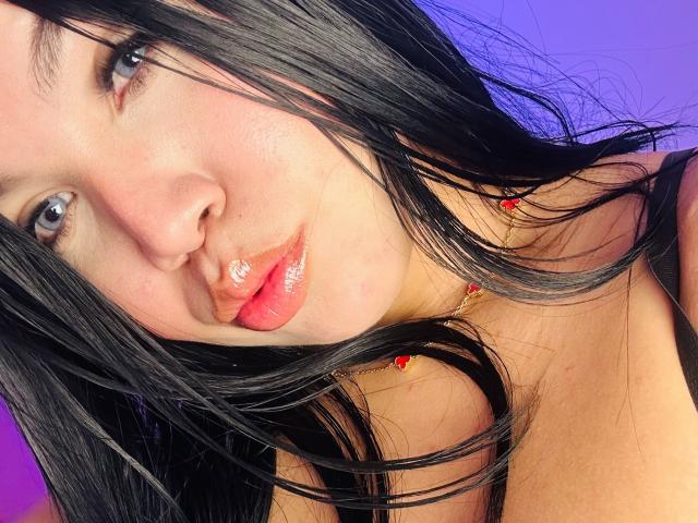 Close-up foto van MiaDenver, een vrouw met bleke huid en zwart haar, die recht in de camera kijkt met glanzende rode lippen. Haar haar bedekt gedeeltelijk haar linker schouder, blauw-paarse achtergrond.