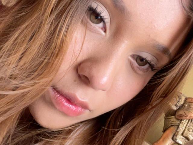 Primer plano del rostro de HeraJuno, con cabello castaño que cubre parcialmente sus ojos, piel clara, labios rosados y pestañas largas. Su expresión es sensual, con un toque de tensión sexual. Centrémonos en su rostro..