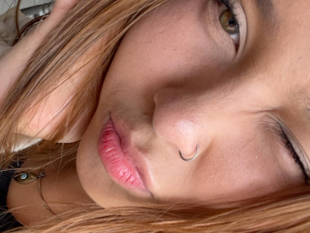 Primo piano di HeraJuno con la pelle abbronzata, gli occhi marroni e le labbra rosa. Ha un anello al naso e il suo viso è parzialmente coperto dai suoi capelli castano chiaro. La sua espressione è seducente..