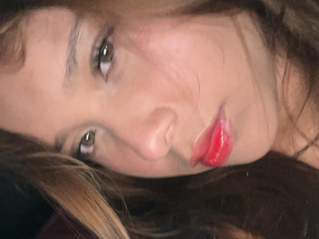 Close-up foto van HeraJuno, gezicht gevuld met zacht licht, bruine ogen die recht vooruit kijken, volle rode lippen die lichtjes open zijn, donker bruin haar met highlights, lichte schaduw onder haar rechteroog, sensuele uitdrukking.