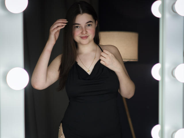 Fotografia di LizabethAmore con la pelle chiara e i capelli lunghi e scuri, che indossa un abito nero senza maniche, in piedi di fronte a lampadine accese, mani che sistemano i capelli, espressione sexy e sicura, sorriso dolce..