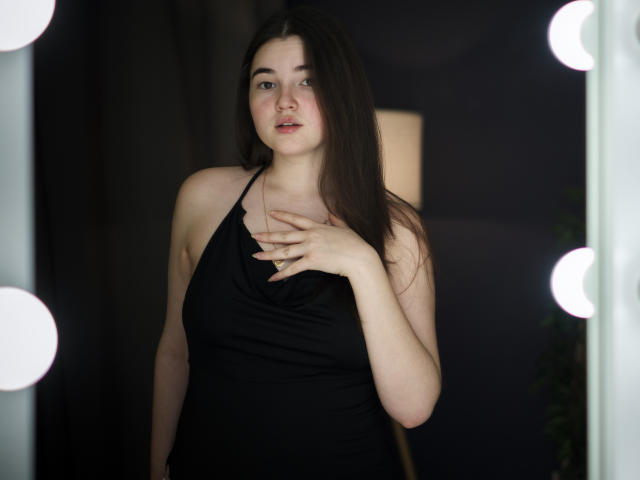 Foto van LizabethAmore met lang donker haar, die een zwarte haltertop draagt, met haar hand op haar borst, staand voor een heldere achtergrond. ronde lampen, die een sterk contrast creëren, intense blik, seksuele spanning.