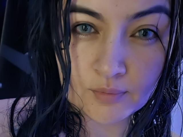 Foto em close-up de LindaSophieX, uma mulher de pele clara, com cabelo escuro e molhado, olhos azuis e pequenas sardas. Ela está olhando diretamente para a câmera com uma expressão ligeiramente neutra. Iluminação azul.