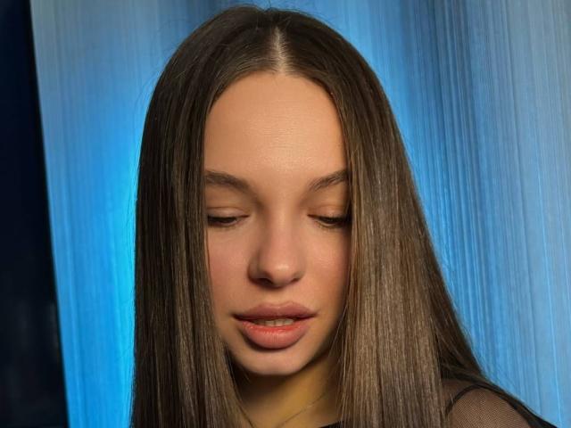 Photograph of CharlenaSable, a young woman with long, straight brown hair, closed eyes, and slightly parted pink lips. Blue gradient background. Her smooth skin is illuminated, creating a sensual atmosphere.