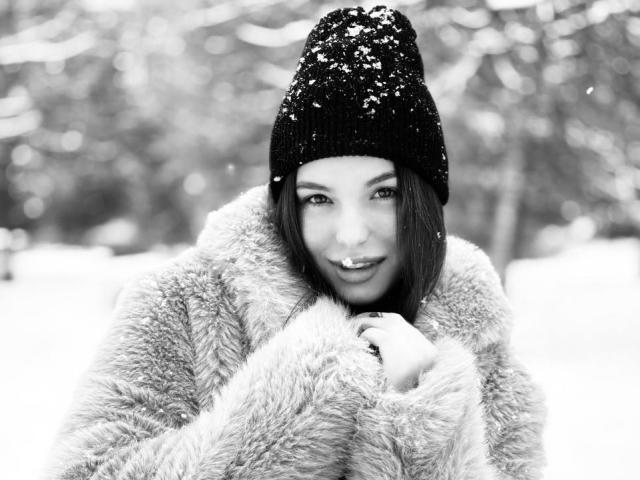 Foto em preto e branco de Charlena Sable, usando um gorro de lã coberto de neve, vestindo um casaco de pele, inclinada para frente, mordendo o lábio. de forma sedutora. Flóculos de neve em seu casaco, fundo nevado borrado. Visual de inverno intenso e sexy..