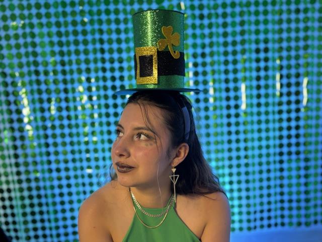 SiaWattson, avec de longs cheveux noirs, portant un haut vert à encolure américaine et un chapeau haut vert scintillant avec un nœud doré. Fond bleu carrelé, regardant sur le côté avec un regard séduisant.