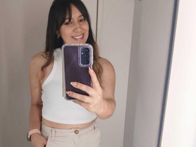 RoxanneBlair tira uma selfie no espelho, sorrindo, com cabelo escuro, vestindo uma blusa branca de alça e calças bege, segurando um objeto preto. telefone com uma capa azul, mão na cintura, tensão sexual.