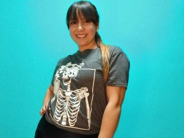 Fotografia de Roxanne Blair com cabelo escuro preso em um rabo de cavalo, vestindo uma camiseta preta com uma estampa de esqueleto branco, em pé. Contra um fundo azul claro, sorrindo e ligeiramente inclinado para frente..