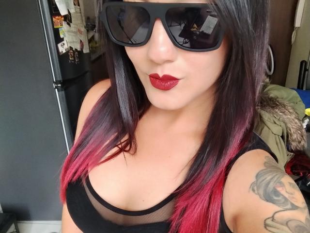 Close-up selfie van MissValentinaRamirez met zwarte zonnebril, rode lipstick en een zwarte kanten top die de decolleté laat zien. Ze heeft donkerbruin haar met roze highlights en een tatoeage op haar linkerarm. Heel erg aantrekkelijk..