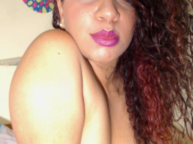 TiaraKitty with curly red-brown hair, purple lipstick, and shiny skin, showing her bare shoulder and upper chest, her tits partially visible, head slightly tilted.