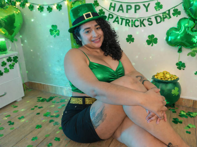 ViickyLee zit op de vloer, met een groene beha, zwarte korte broek en een groene klavertje vier-hoed, en glimlacht naar de camera. Groene klavertjes en ballonnen, een pot met goud op de achtergrond.