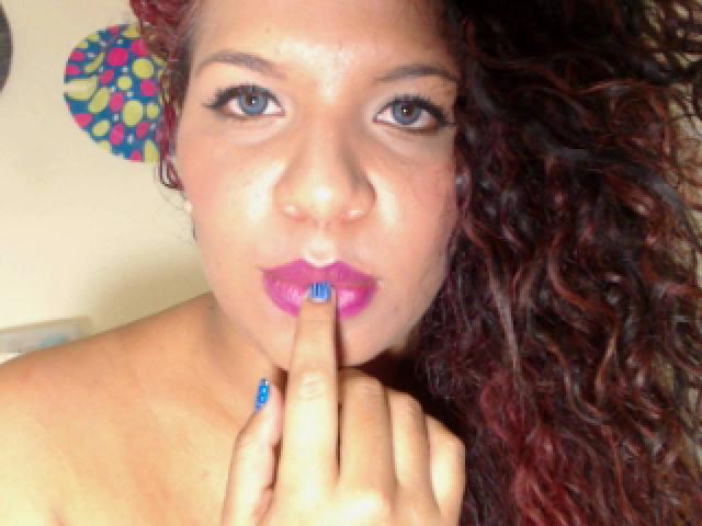 TiaraKitty with curly red-brown hair, blue nail polish, pink lipstick, and finger in mouth, showing off her shiny skin and blue eyes. Boobs not visible, focus on face.