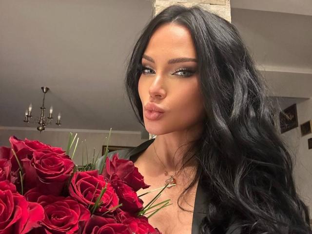 Photograph of BrookeEmber with long black hair, pouty lips, and heavy makeup, holding a bouquet of vibrant red roses. She has a seductive gaze and a black top. Chandelier in the background.