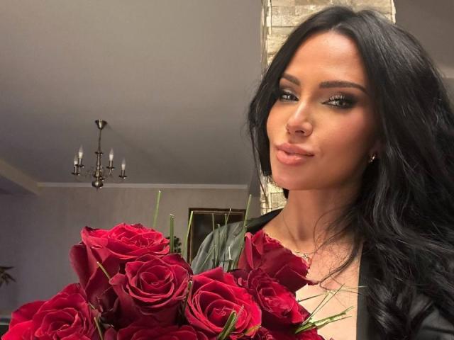 Photographie de BrookeEmber avec de longs cheveux noirs, tenant un bouquet de roses rouges, un anneau au nez et un maquillage des yeux aux tons fumés, regardant directement face à la caméra, avec une expression légèrement boudeuse..