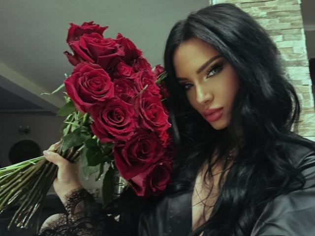 Fotografia di BrookeEmber con lunghi capelli neri, che tiene un bouquet di rose rosse. Indossa una camicetta nera in pizzo e guarda direttamente l'obiettivo con uno sguardo seducente. Lo sfondo include un muro di mattoni..