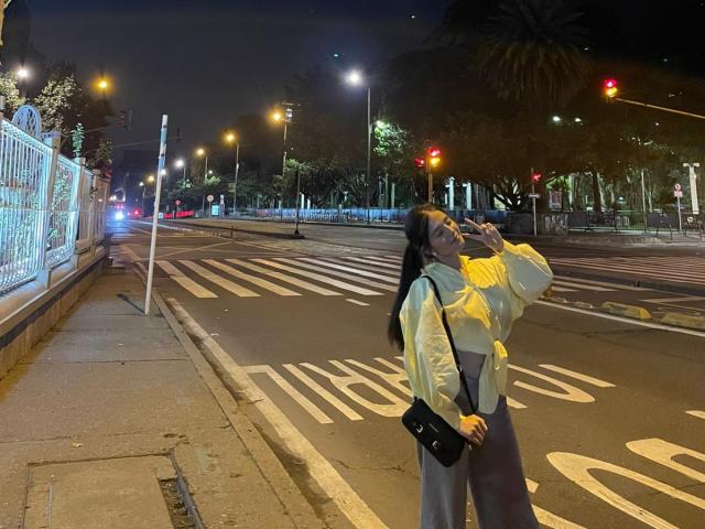 Nachtaufnahme von Ameliiex, die an einer Straßenecke steht, eine gelbe Jacke, graue Hosen und eine schwarze Umhängetasche trägt. Ameliiex blickt auf, im Hintergrund sind Lichter und Straßenmarkierungen zu sehen. Verdammt sexy, urbaner Vibe..