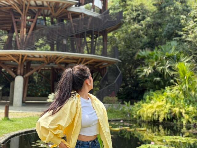 Ameliiex stands in profile, wearing a yellow jacket over a white crop top, looking away with her hand on her hip. Background shows a modern building and lush greenery. Sunlight highlights her long dark hair.