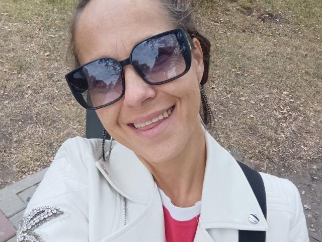 Primer plano de Liora Weiss sonriendo, con gafas de sol negras grandes, una chaqueta blanca y una camisa roja. Tiene una figura delgada, piel clara y cabello castaño recogido. Hierba en el fondo. Qué lindo..