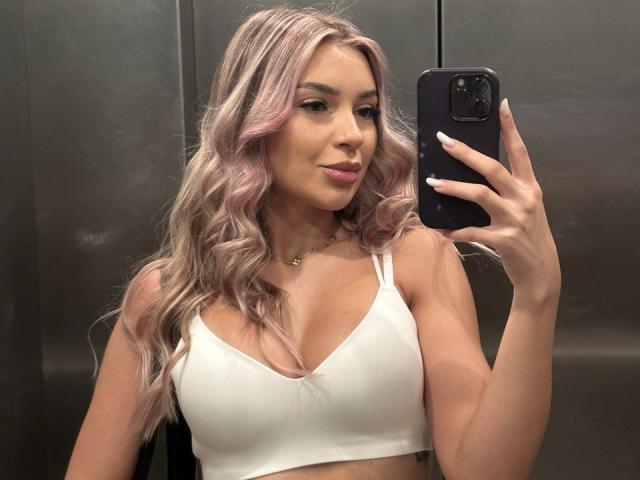 Photograph of ArtTeasee, a sexy woman with long, wavy, pinkish-blonde hair, wearing a white sports bra, holding a black smartphone, taking a mirror selfie. She has a confident, sexy expression.