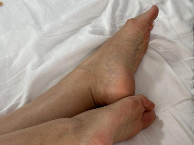 Close-up of ArtTeasee's bare feet with slightly dirty soles, crossed at the ankles on a crumpled white sheet. Skin shows natural textures and shadows, conveying a relaxed, intimate moment.