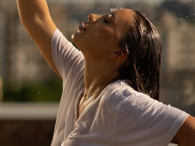 Photograph of ArtTeasee, a wet woman with dark hair, arms raised, white shirt clinging to her body, sunlight highlighting droplets, cityscape blurred in the background, intense, sexual tension.