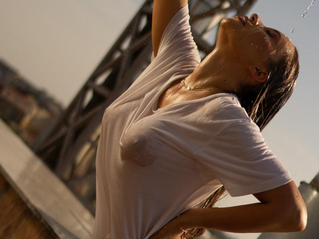 ArtTeesee with wet, white t-shirt clinging to her body, nipples visible. She arches back, head tilted up, water droplets on her skin. Sunlight highlights her wet hair and toned physique.